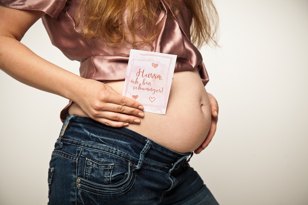 Schwangere Frau hält ein Schild vor ihren Bauch mit der Aufschrift "Hurra, ich bin schwanger!"